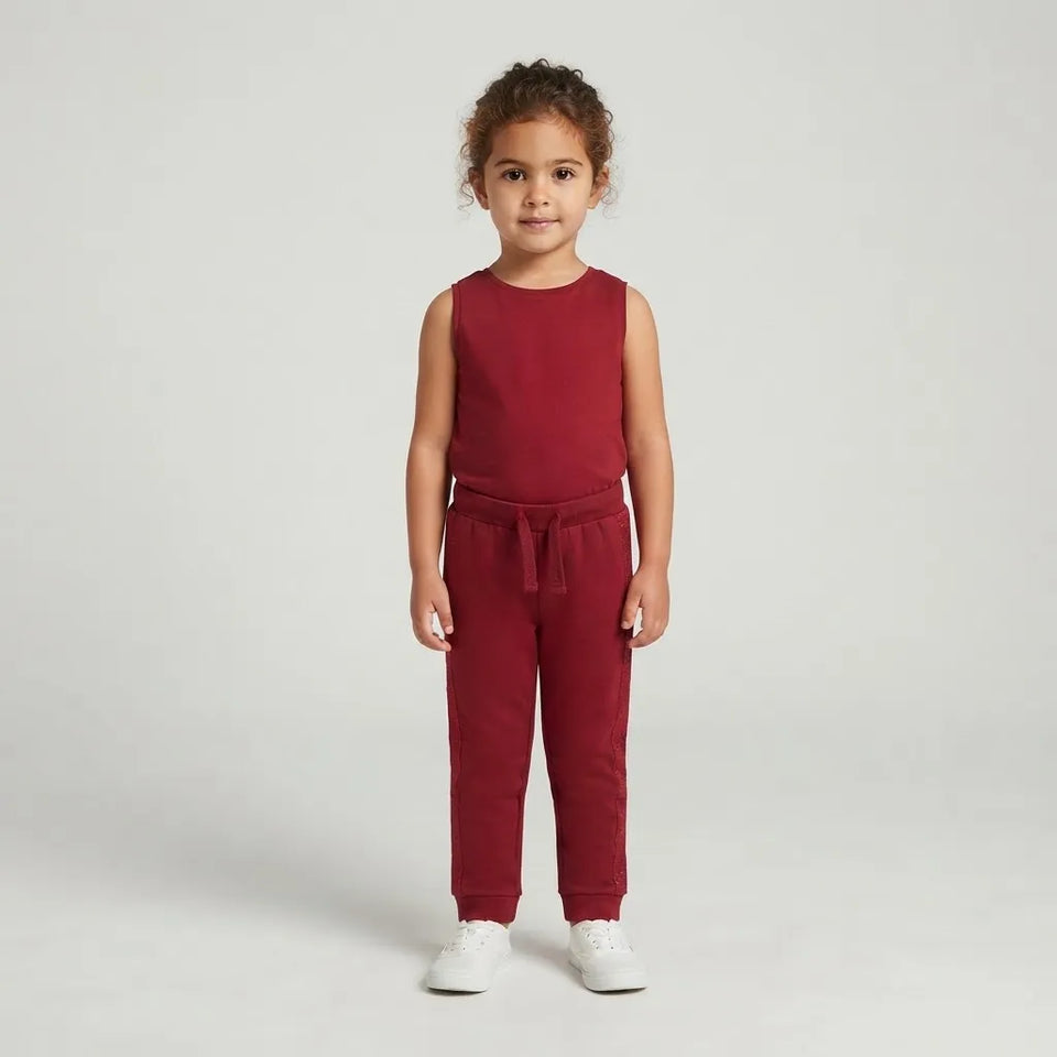 A young child stands against a light gray studio background wearing a sleeveless red top and matching red jogger-style pants with white sneakers.