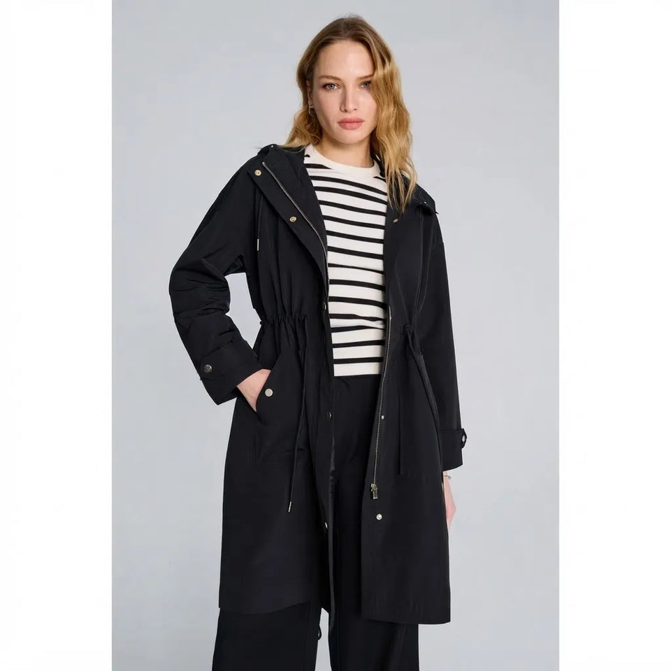 A woman modeling a black hooded trench coat over a black and white striped top and black pants, photographed against a light gray studio background.