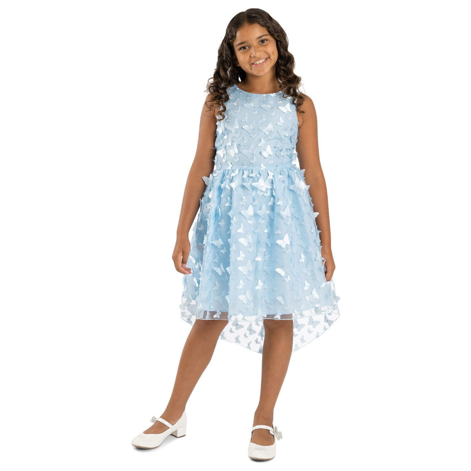 A young girl modeling a sleeveless light blue dress with a textured bodice and a layered tulle skirt decorated with white butterfly appliqués, paired with white Mary Jane shoes, on a solid white background.