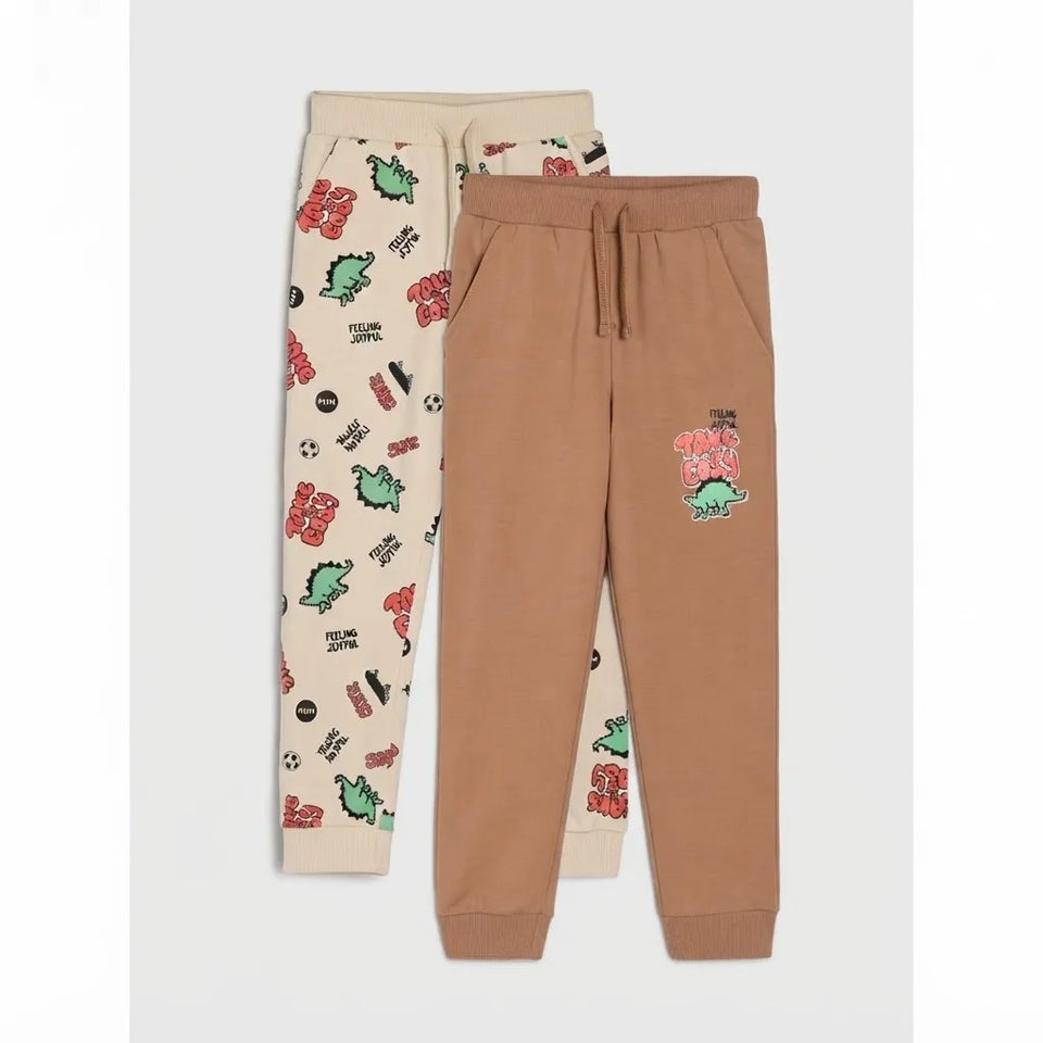 Two pairs of kids' sweatpants laid flat on a light gray background. The front pair is tan with a small colorful dinosaur graphic on the left thigh.