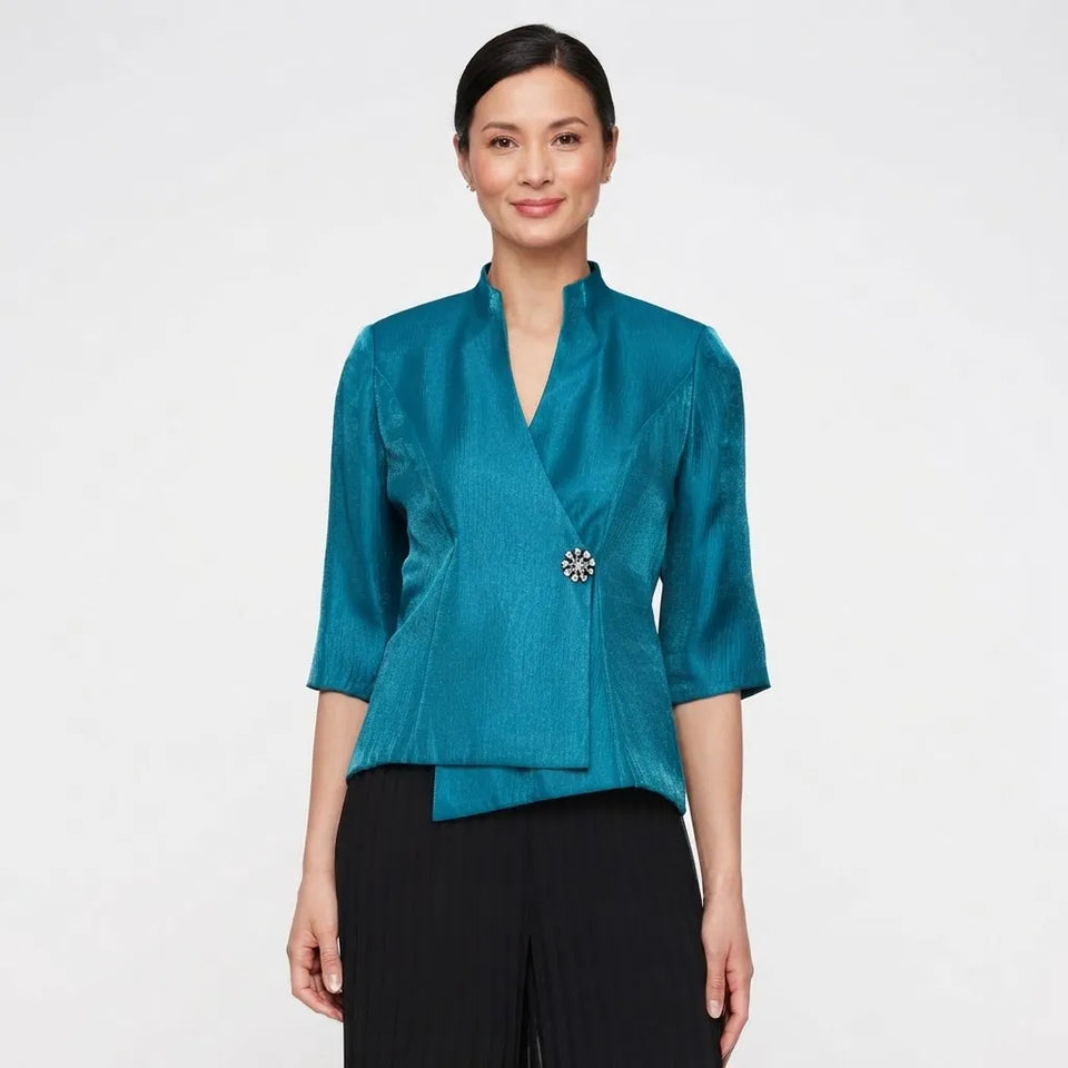 A woman modeling a teal textured wrap-style blouse with three-quarter sleeves and a decorative brooch at the waist, paired with black pleated pants, against a plain light gray background.