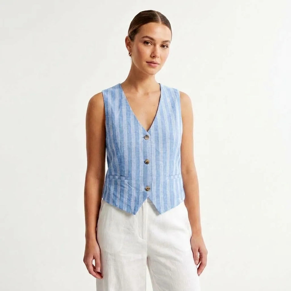 A woman modeling a sleeveless light blue and white vertically striped vest with three brown buttons, paired with white pants, against a plain white studio background.