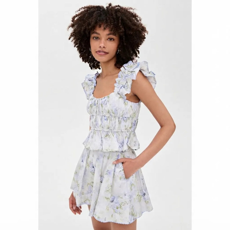 A woman modeling a sleeveless ruffled mini dress with a smocked bodice and a light blue and green floral print on a white background, set against a plain light gray studio backdrop.