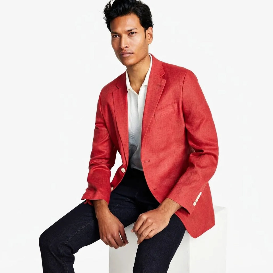 A man wearing a textured red blazer over a white collared shirt and dark jeans, seated on a white block against a plain white background.