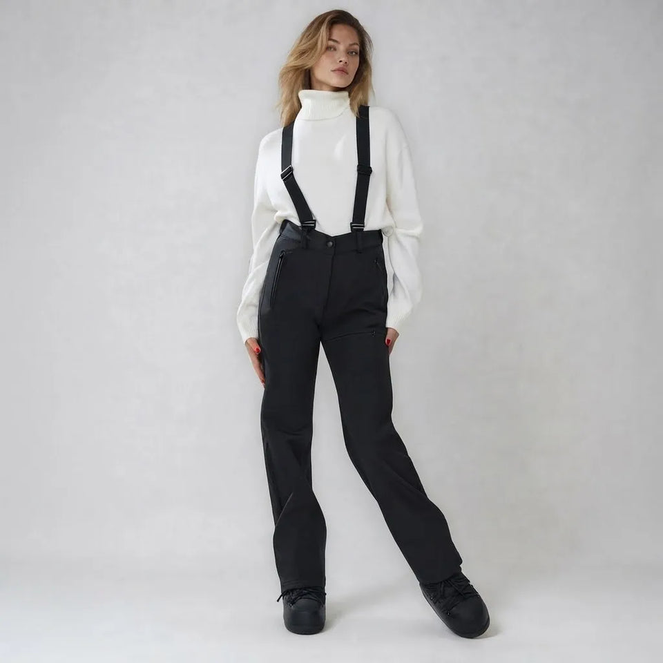 A woman modeling black wide-leg ski pants with black suspenders, paired with a white turtleneck sweater and black sneakers, against a light gray studio background.