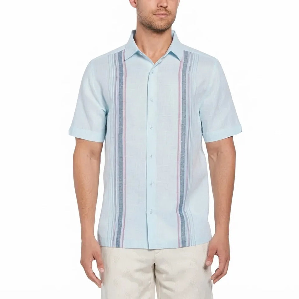 A man wearing a light blue short-sleeve guayabera-style shirt with vertical stripes in blue, pink, and white, paired with beige pants, against a plain white background.