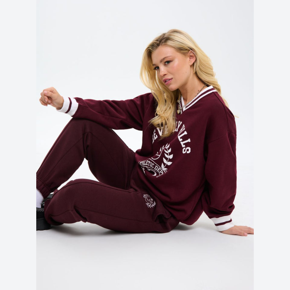 A woman sitting on a light gray studio floor wearing a matching burgundy sweatshirt and jogger pants set with white varsity-style trim and lettering.
