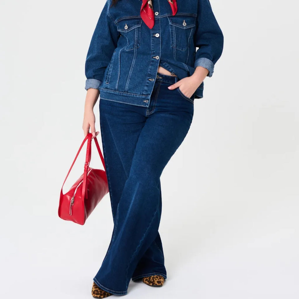 A model wearing a dark blue denim jacket and matching flare jeans, styled with a red neck scarf, leopard print shoes, and carrying a red handbag against a plain white background.