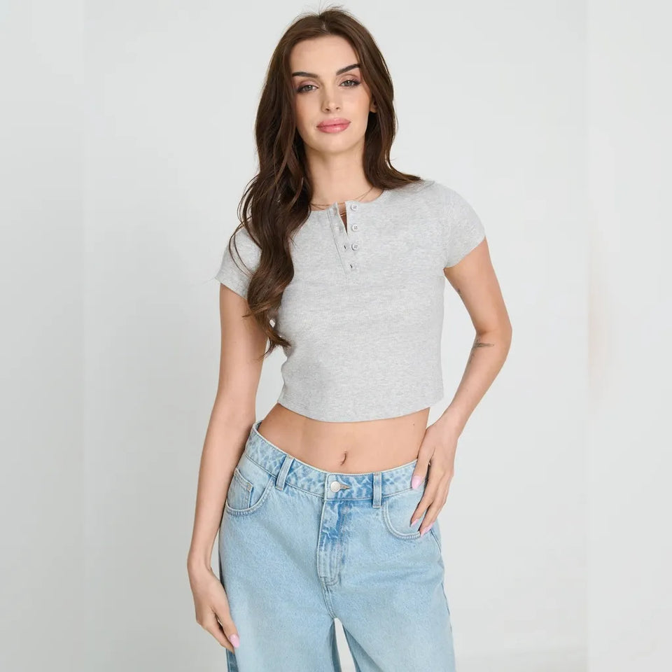 A woman modeling a light gray short-sleeve crop top with a split neckline and button detail, paired with light blue jeans, against a plain light gray studio background.