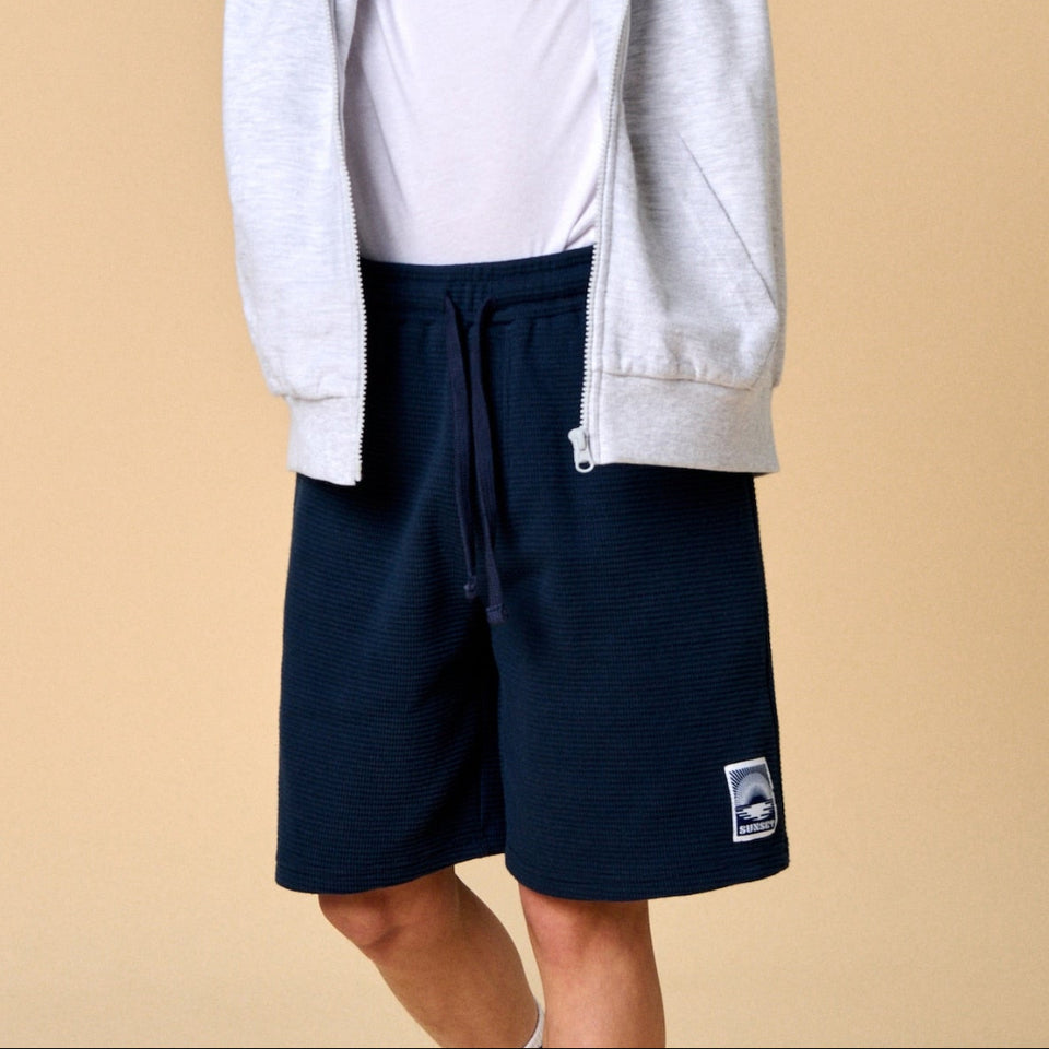 A child model wearing a light gray zip-up hoodie over a white t-shirt and navy blue sweat shorts, posing against a solid beige studio background.
