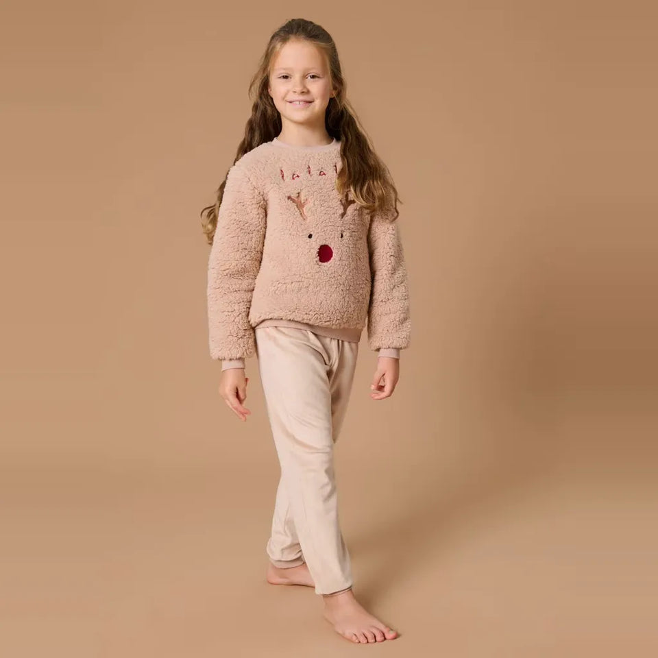 A young girl stands barefoot against a solid beige studio background, wearing a fuzzy light pink long-sleeve sweater with a reindeer face graphic and light beige pants.