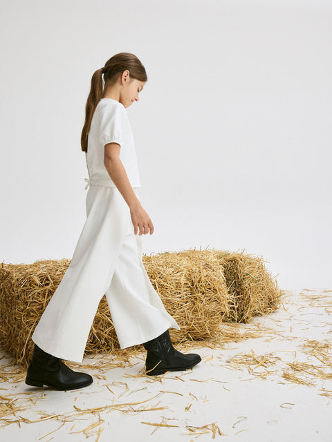 Person wearing a white outfit and black boots standing on hay bales against a white background