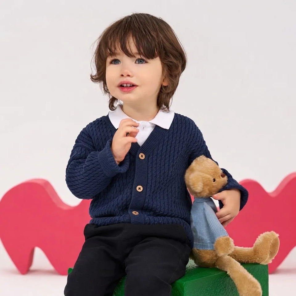 A young child with brown hair sits on a green block wearing a navy blue cable-knit cardigan over a white collared shirt, paired with black pants. The child is holding a brown teddy bear and is posed against a white background with pink heart-shaped props.