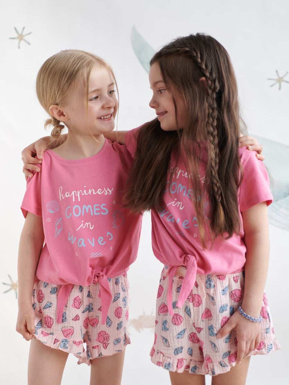 Two young girls wearing matching pink outfits with text and floral skirts on a white background.
