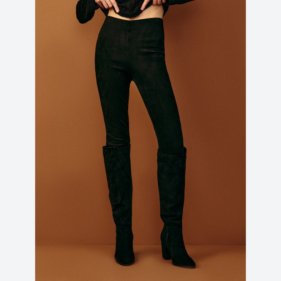 Black high-waisted skinny pants with a matte suede-like finish, shown on a model from the waist down against a solid warm brown studio background.