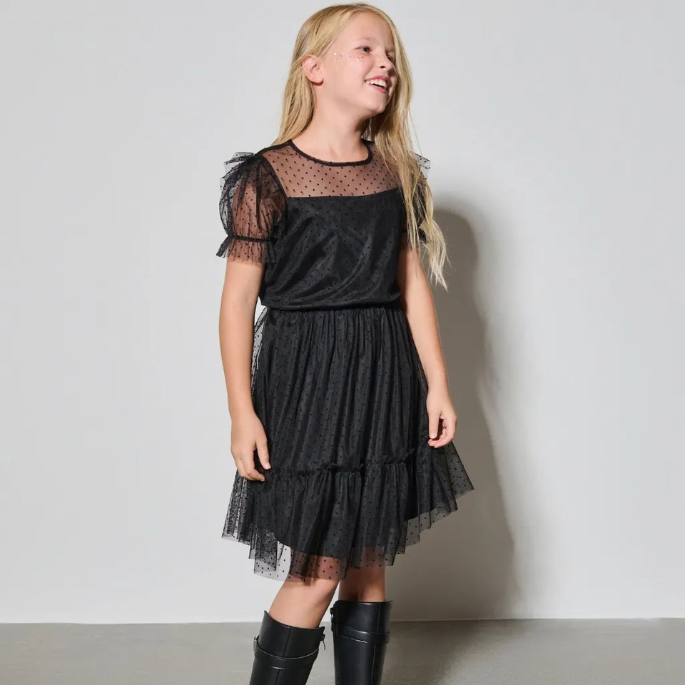 A young girl with blonde hair wearing a black tiered tulle dress with sheer mesh sleeves and black knee-high boots, standing against a light gray studio wall.