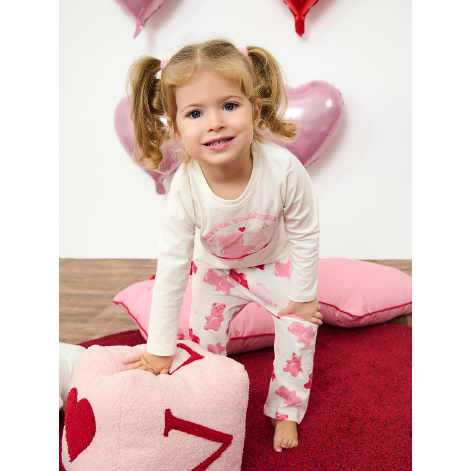 A young girl with blonde pigtails sits on a red carpet wearing a white long-sleeve pajama top and matching white pajama pants with pink teddy bear and heart patterns. She is posing with pink heart-shaped pillows and a plush pink pillow in the foreground.