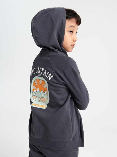 Child wearing a dark gray hoodie with a mountain graphic and text on the back against a white background