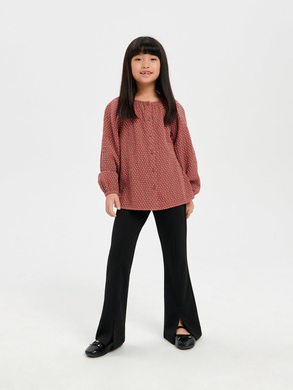 Young girl wearing a pink textured sweater and black pants on a white background