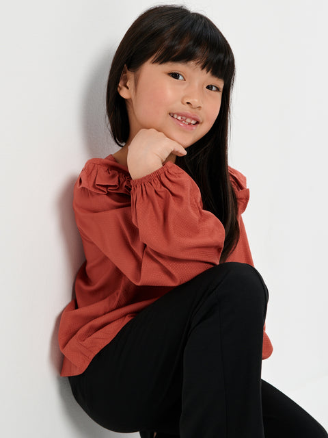 Young girl wearing a rust-colored blouse and black pants against a white background