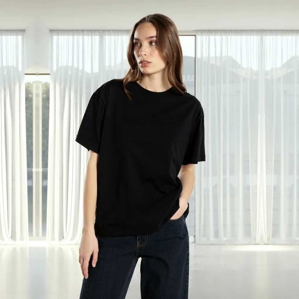 A woman wearing a plain black short-sleeve crewneck t-shirt and dark blue jeans, standing in a bright room with sheer white curtains and a blurred mountain background.