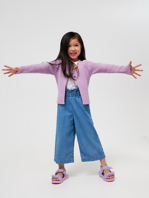 Child wearing a pink cardigan, white shirt, blue jeans, and purple sandals on a white background