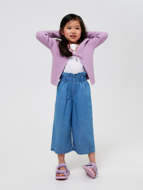 Young girl wearing a pink cardigan, white shirt, blue jeans, and purple sandals on a white background