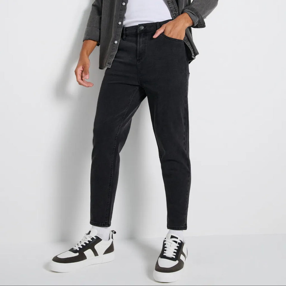 A person wearing slim-fit black jeans, styled with a gray button-up shirt, white t-shirt, and black-and-white sneakers against a light gray studio background.