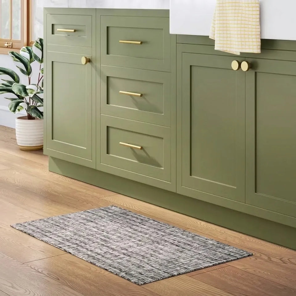 An olive green kitchen vanity cabinet set with brass hardware, shown in a styled kitchen setting with a gray textured bath mat on a wood floor, a potted plant, and a checkered towel.
