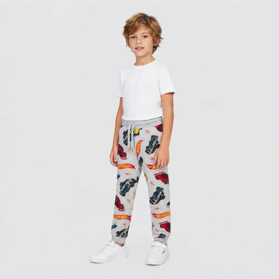 A young boy modeling light gray sweatpants with a colorful Hot Wheels car print, paired with a white t-shirt and white sneakers, against a plain light gray background.