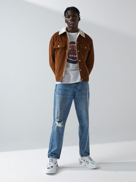 Person wearing a brown jacket, white shirt with a pattern, blue jeans, and white sneakers on a white background