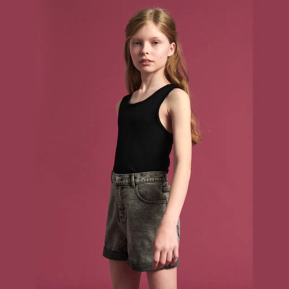 A young girl with long light brown hair stands against a solid dusty rose background, wearing a black sleeveless tank top and acid-wash gray denim shorts.