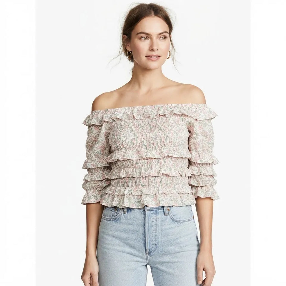 A woman wearing a light pink and white ruffled off-the-shoulder top with three-quarter sleeves, paired with light-wash blue jeans, against a plain white background.