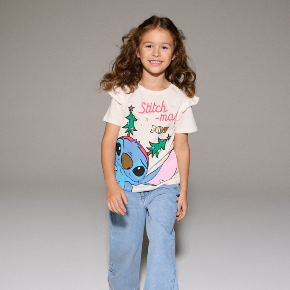 A young girl with long brown hair stands against a neutral gray studio background wearing a white graphic t-shirt with a blue cartoon character and the text 'Stitch - may DOO!', light blue wide-leg jeans, and pink sneakers.