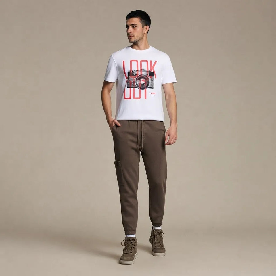 A man modeling a white crewneck t-shirt with a red and black graphic print that reads 'LOOK OUT' with a camera illustration, paired with brown jogger pants and brown sneakers, against a solid beige studio background.