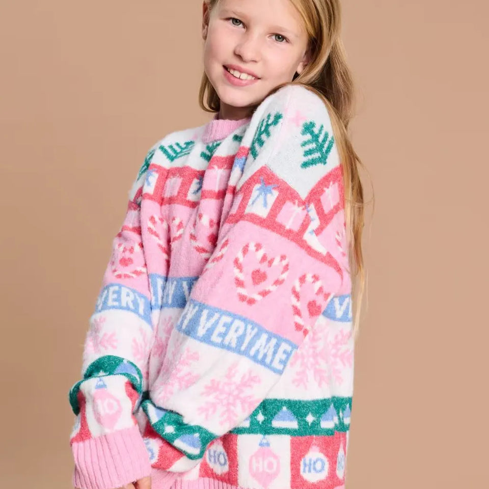 A young girl with blonde hair smiling while wearing a pink and white holiday-themed knit sweater with green and blue accents, set against a solid beige background.