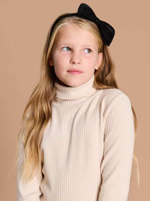 Young girl with long blonde hair wearing a beige turtleneck sweater against a beige background