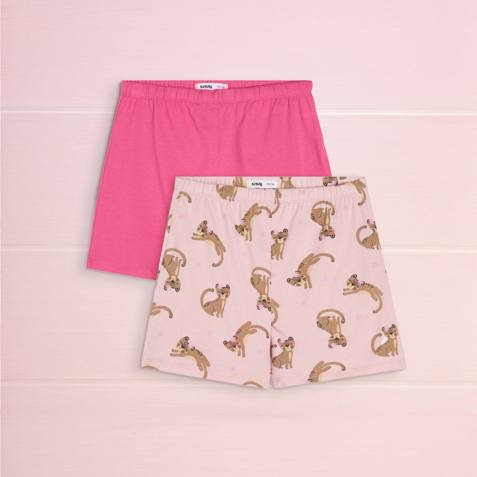 Two pairs of children's boxer shorts laid flat on a pale pink background. The back pair is solid bright pink, and the front pair is light pink with an allover print of playful brown cats.