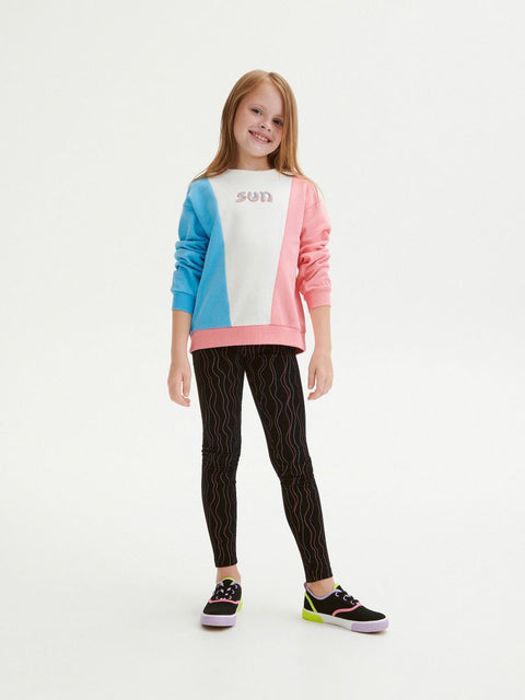 Young girl wearing a colorful sweater and black patterned leggings on a white background