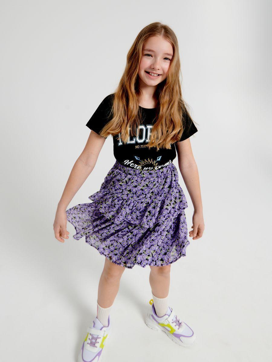 Young girl wearing a black t-shirt with text and a purple floral skirt on a white background