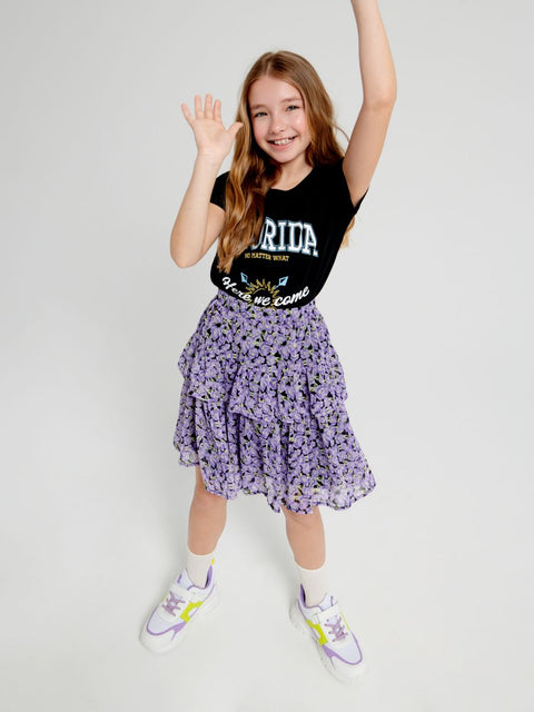 Young girl wearing a black top with text and a purple floral skirt on a white background