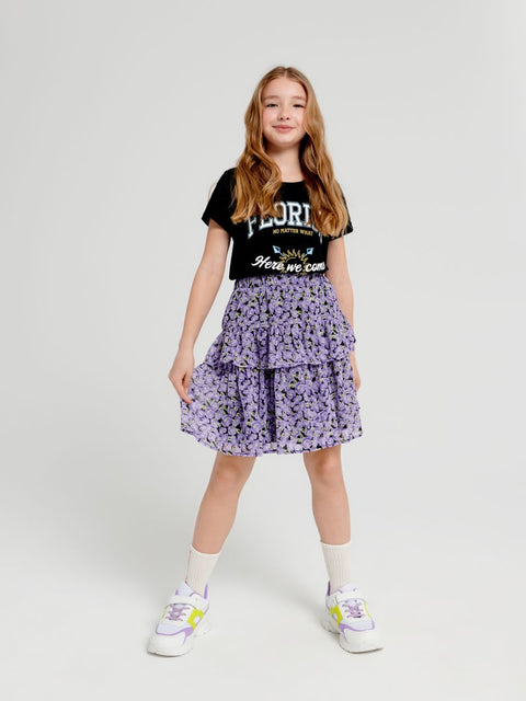 Young girl wearing a black t-shirt with text and a purple floral skirt on a white background