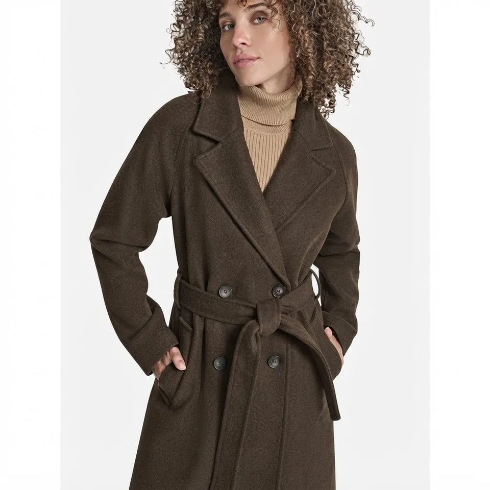 A woman wearing a dark brown double-breasted wool coat with a matching waist tie belt, styled over a beige ribbed turtleneck sweater, set against a plain light gray background.