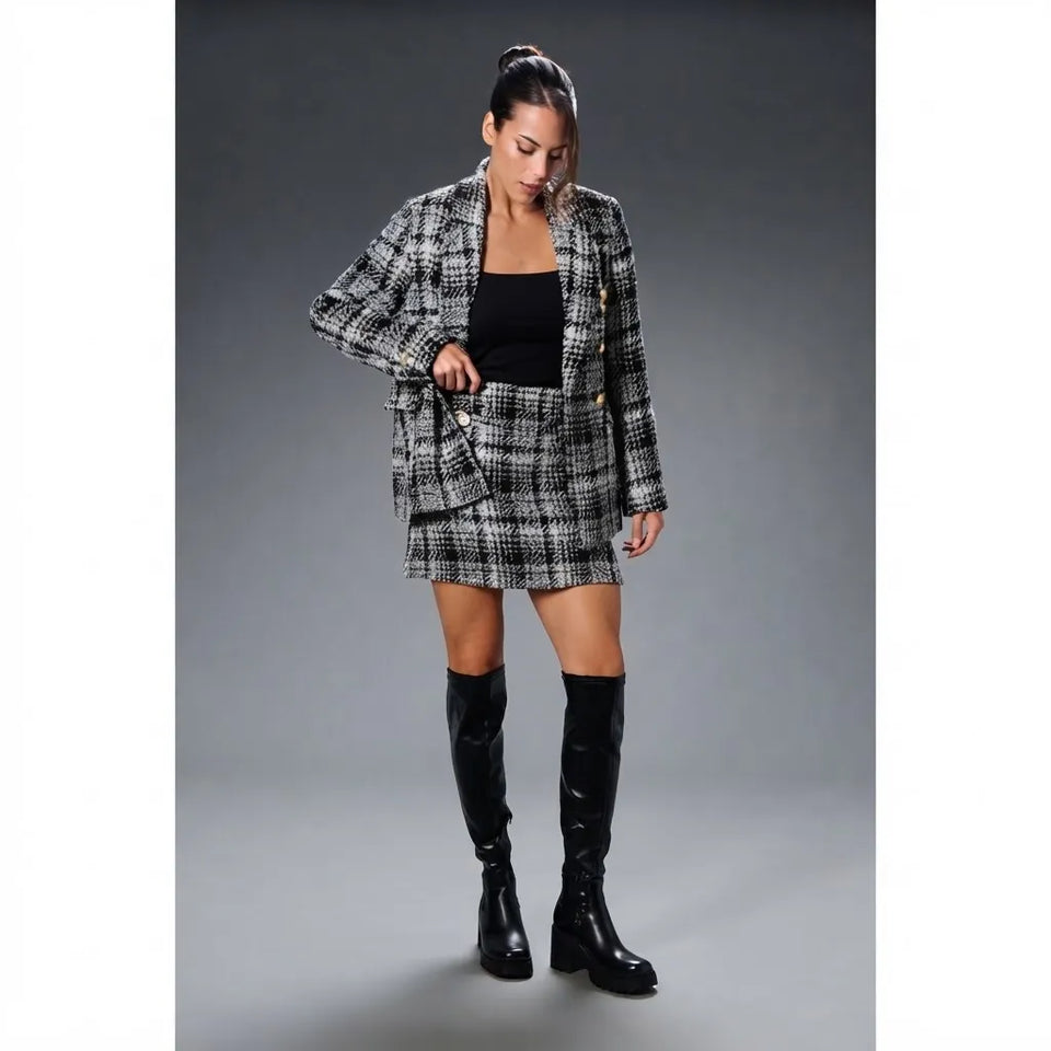 A woman models a black and white plaid blazer and matching mini skirt set, styled with a black top and black over-the-knee boots, against a dark gray studio background.