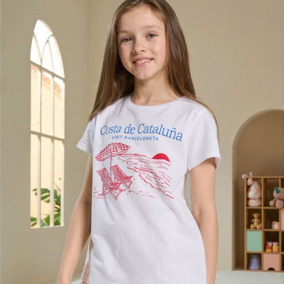 A young girl wearing a white graphic t-shirt with 'Costa de Catalunya' printed in blue and red, paired with beige pinstriped shorts, standing in a brightly lit room with arched windows.