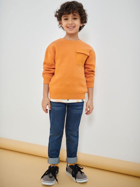 Child wearing an orange sweater and blue jeans standing against a white wall.