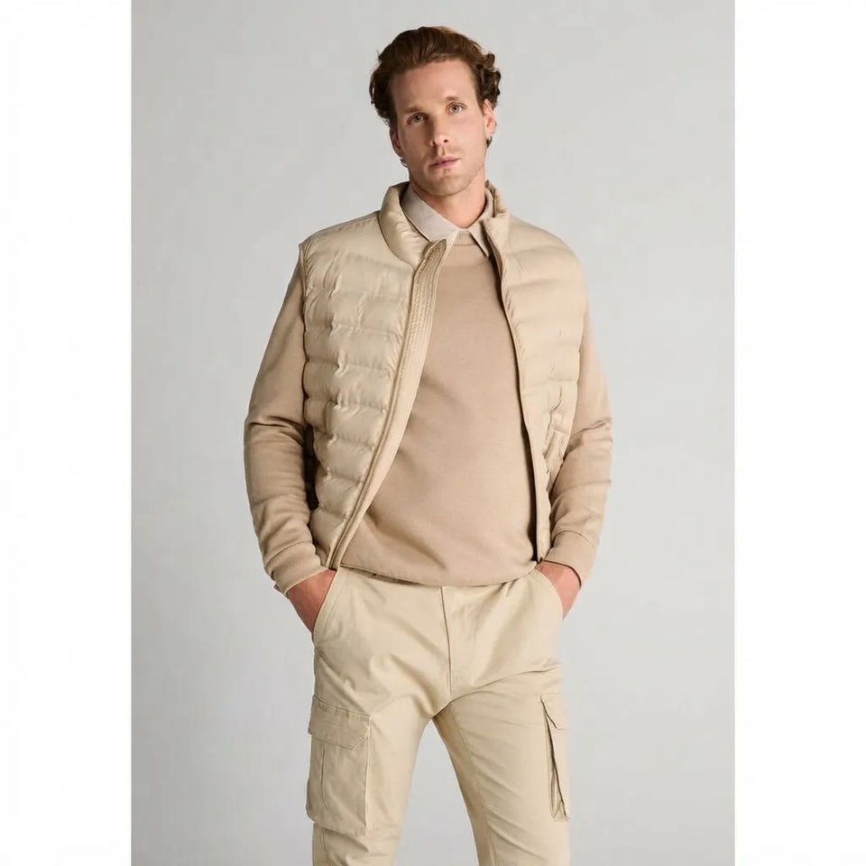 A man wearing a beige quilted puffer vest over a matching beige sweater and light gray collared shirt, paired with beige cargo pants, standing against a light gray studio background.
