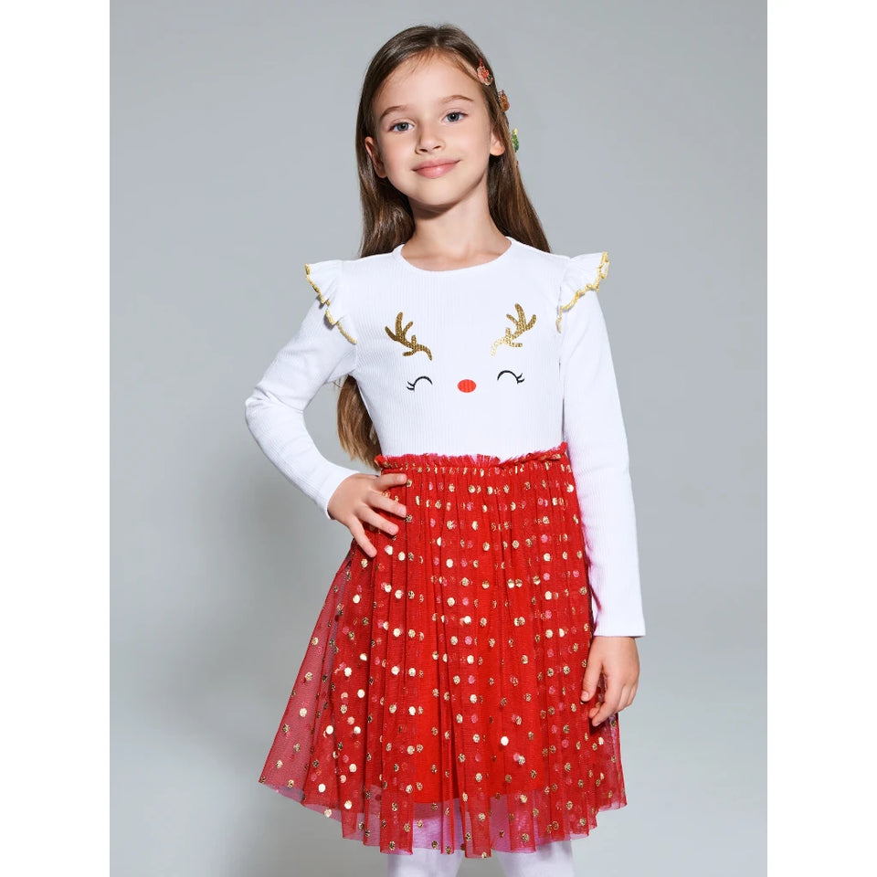 A young girl modeling a festive holiday outfit consisting of a white long-sleeve top with gold reindeer antlers and a red tulle skirt with gold polka dots, set against a plain gray studio background.