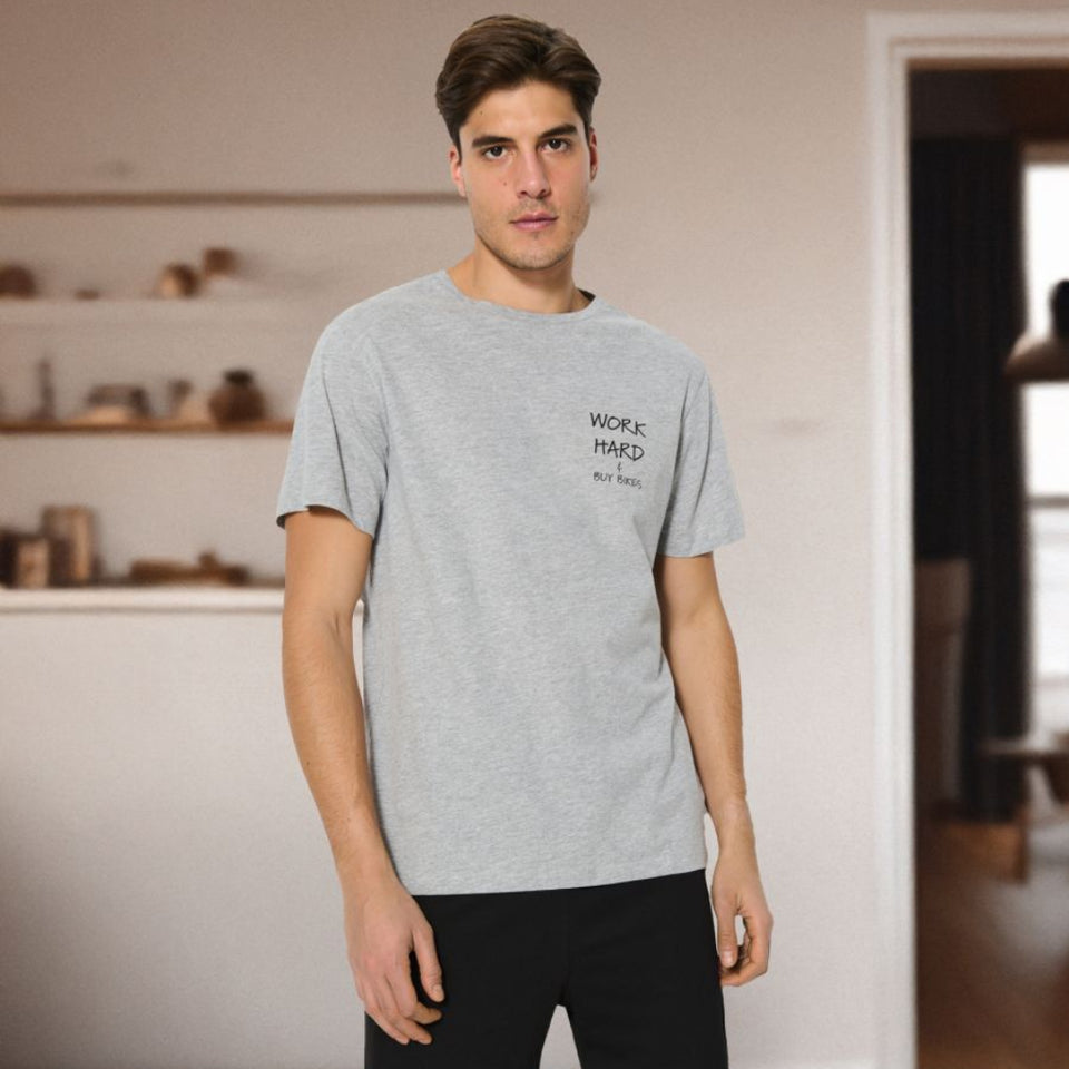 A man wearing a light gray heather crewneck t-shirt with a small black graphic on the left chest and black shorts, standing in a modern, brightly lit room with wooden floors.