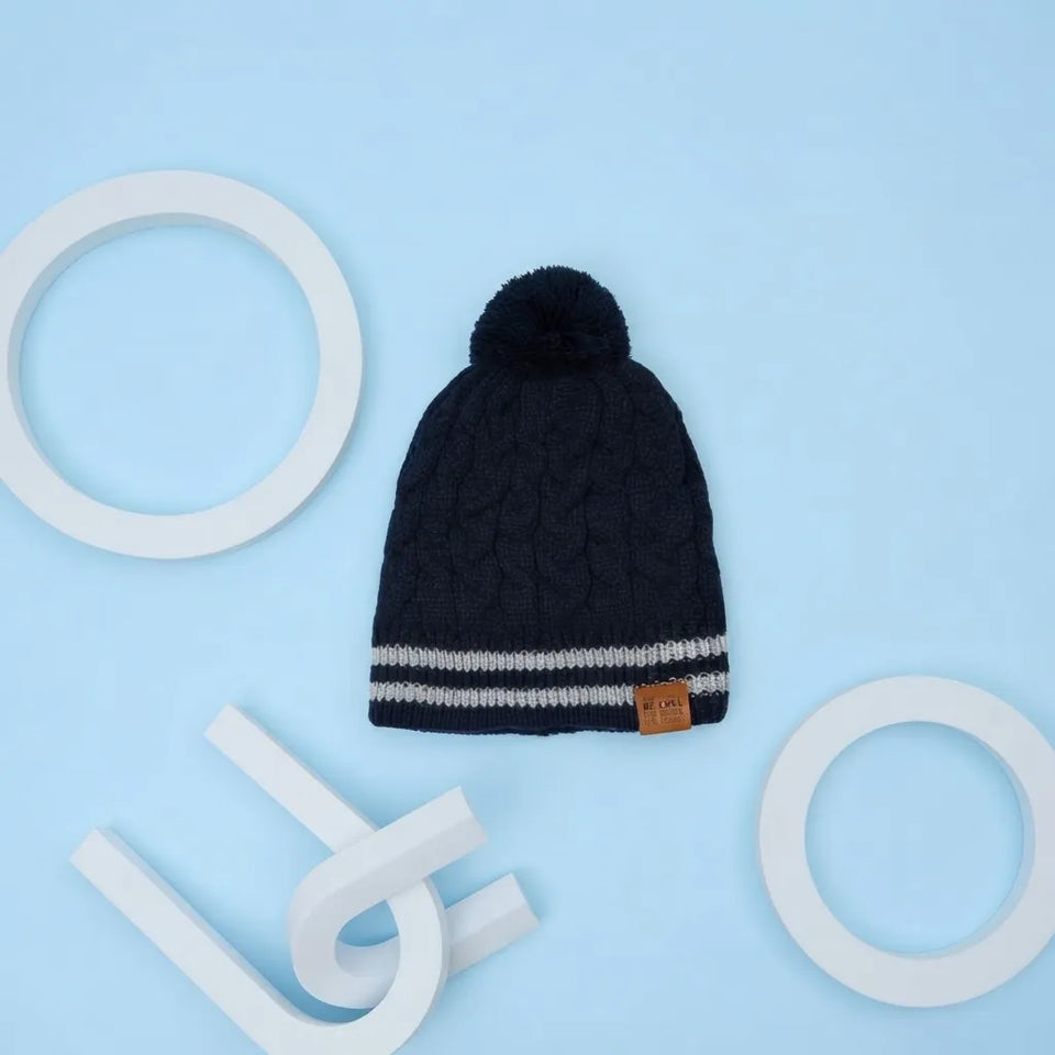 A dark navy blue cable-knit beanie with a matching pom-pom, featuring two horizontal white stripes near the brim and a small brown leather brand tag. The beanie is displayed on a light blue background with white geometric props.